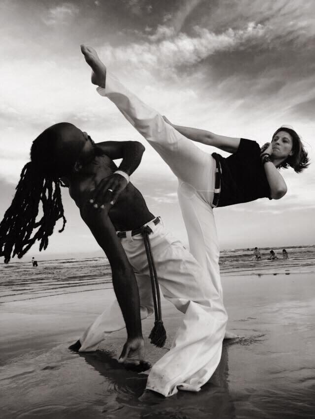 woman in a high kick and man dodging on a beach in a game of capoeira
