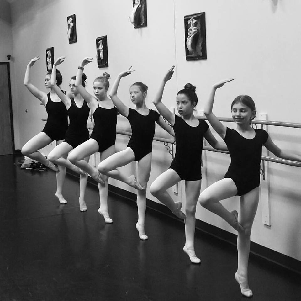 a group of young women in black leotards balancing on one foot at the barre