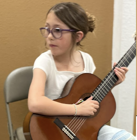 9 year old girl with brown hair playing guitar, looking to her right.